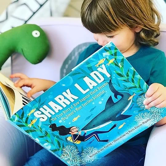 Child reading a colorful children's book titled 'Shark Lady' with an illustration of a shark and diver on the cover, next to a green plush toy resembling a dinosaur.