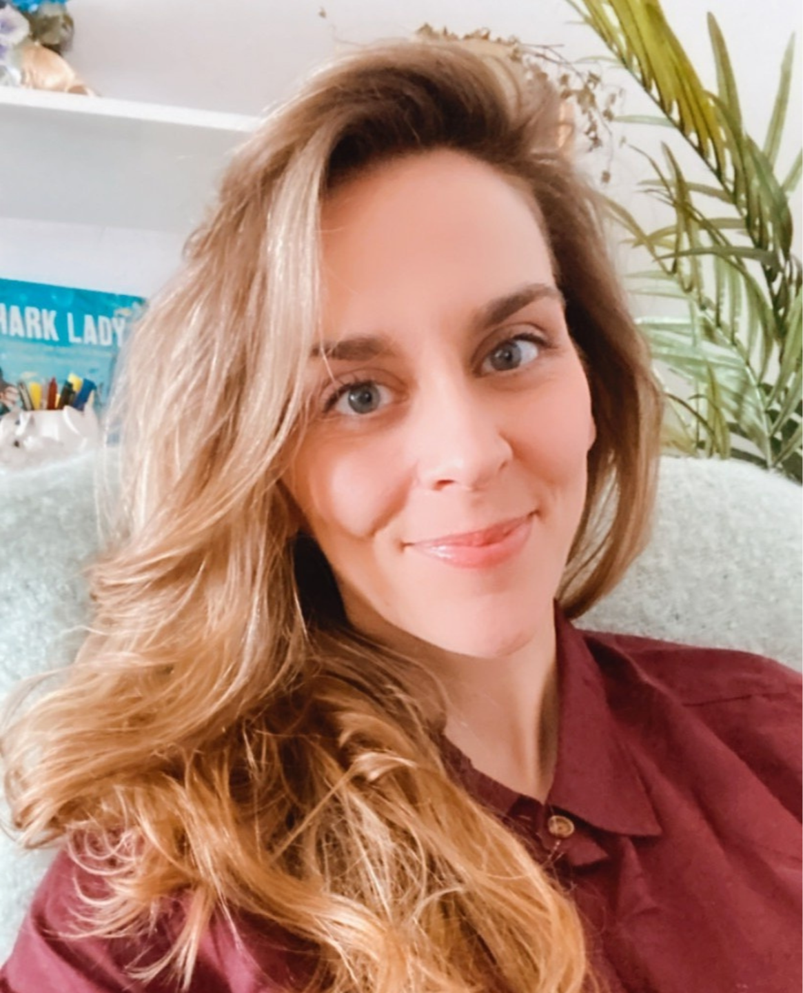 Close-up of a woman with long, wavy brown hair and blue eyes, smiling, wearing a burgundy shirt, sitting in a room with a plant and a bookshelf in the background.