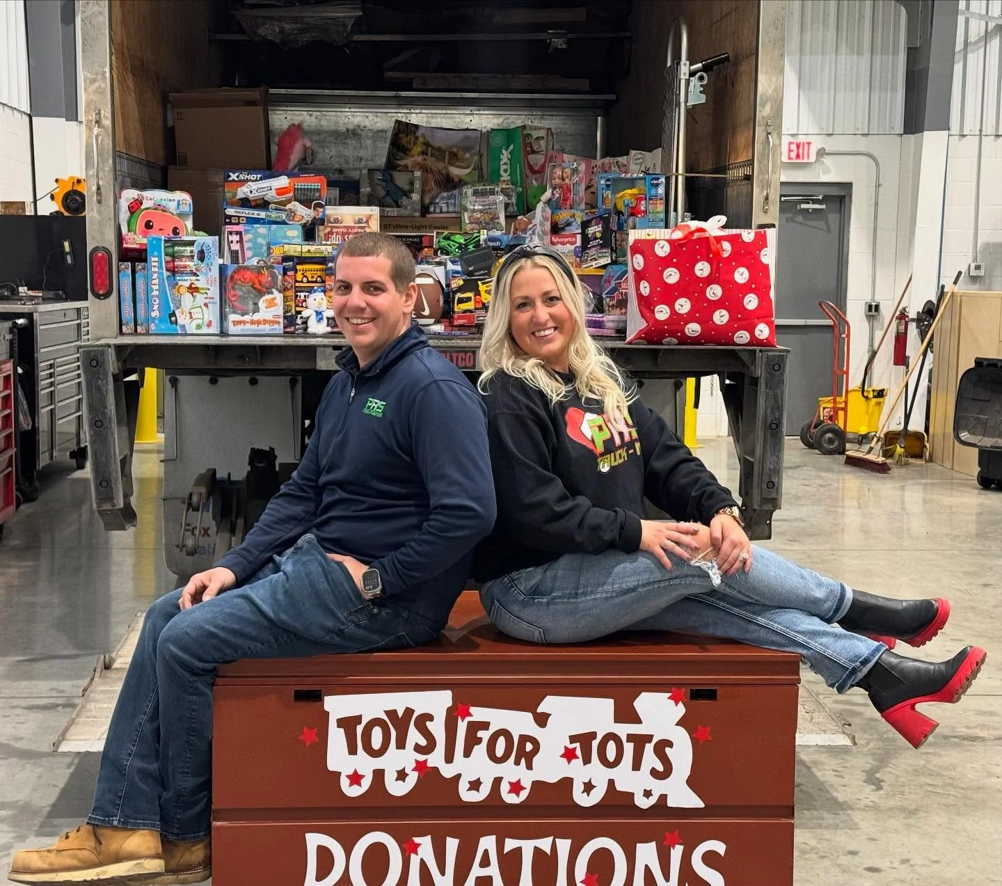 Two smiling people, a man and a woman, sitting back-to-back on a donation box labeled 'TOYS FOR TOTS DONATIONS,' with a large collection of toys in the truck behind them.