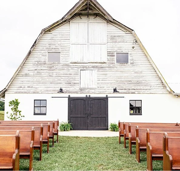 A rustic barn converted into an event space with black double doors, surrounded by wooden pews and green grass, with small trees and windows.