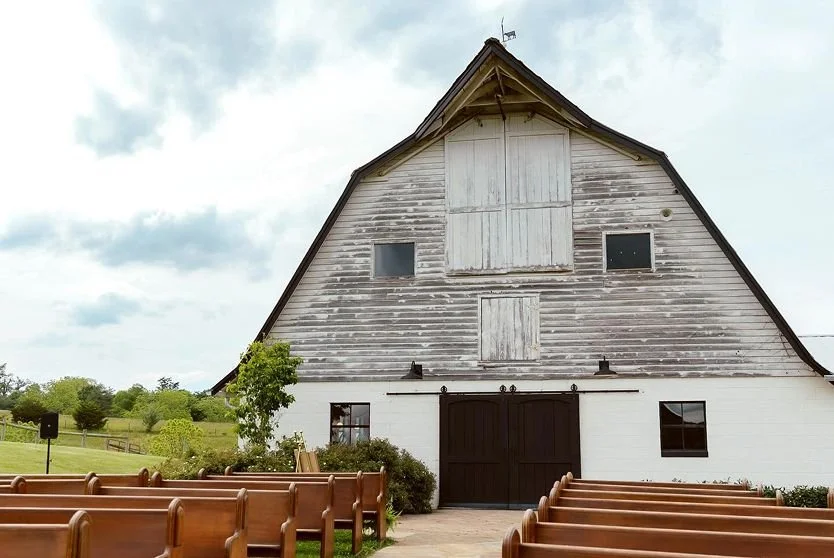 A rustic barn with weathered wood siding, large sliding doors, small windows, and a split-shake roof. There are wooden pews in front of the barn, and nearby greenery and a cloudy sky.