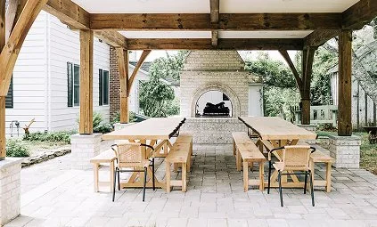 Outdoor patio with wooden pergola, dining tables, chairs, and a stone fireplace in a backyard setting.