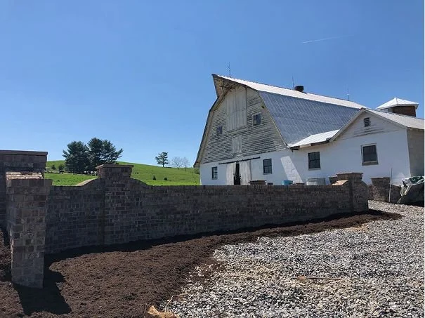 A white barn with a metal roof on a hill, surrounded by a brick foundation and green fields, under a clear blue sky.