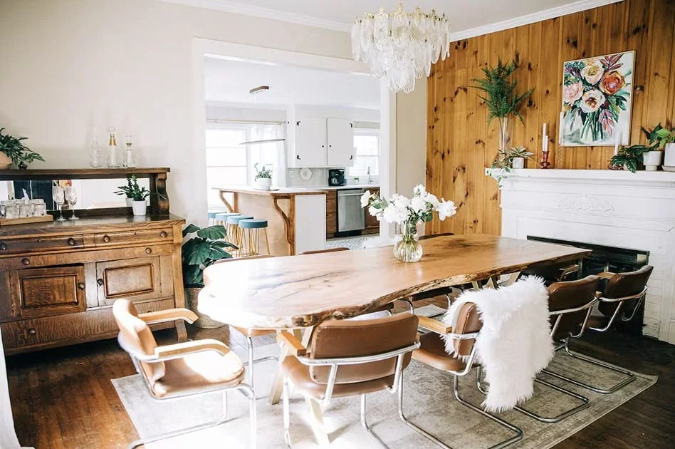 Dining room with a large wooden table, six leather chairs, and a white sheepskin throw. The background includes a wood-paneled wall with a floral painting, a white fireplace mantel, potted plants, and a chandelier overhead.
