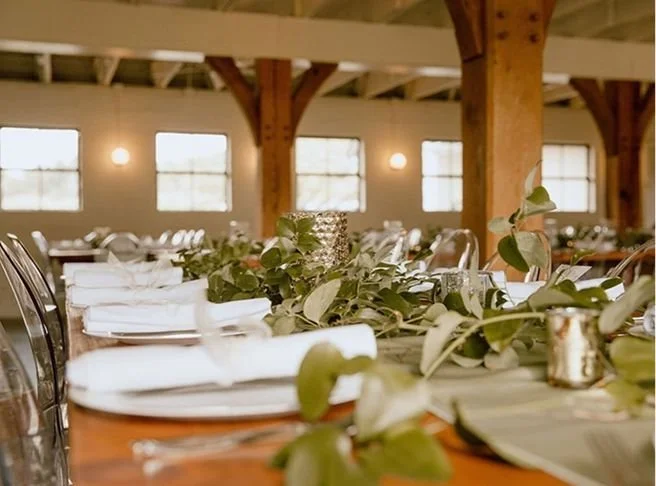 A decorated banquet table with white plates, silverware, green foliage, and candles in a rustic event space with exposed wooden beams and windows.