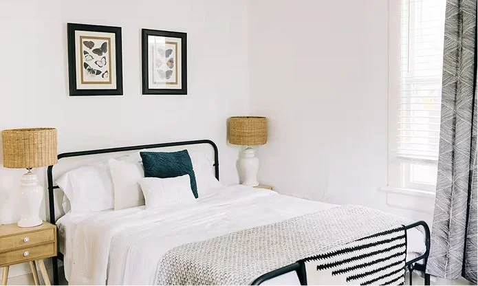 A bedroom with a black metal bed frame, white bedding, and a dark green pillow. Two nightstands with white ceramic lamps and woven rattan lampshades. Wall art featuring botanical prints, a window with white blinds and patterned curtains.