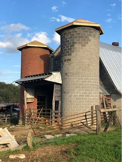 A rustic farm building with two silo towers, one red brick and one gray concrete, under a blue sky with clouds.