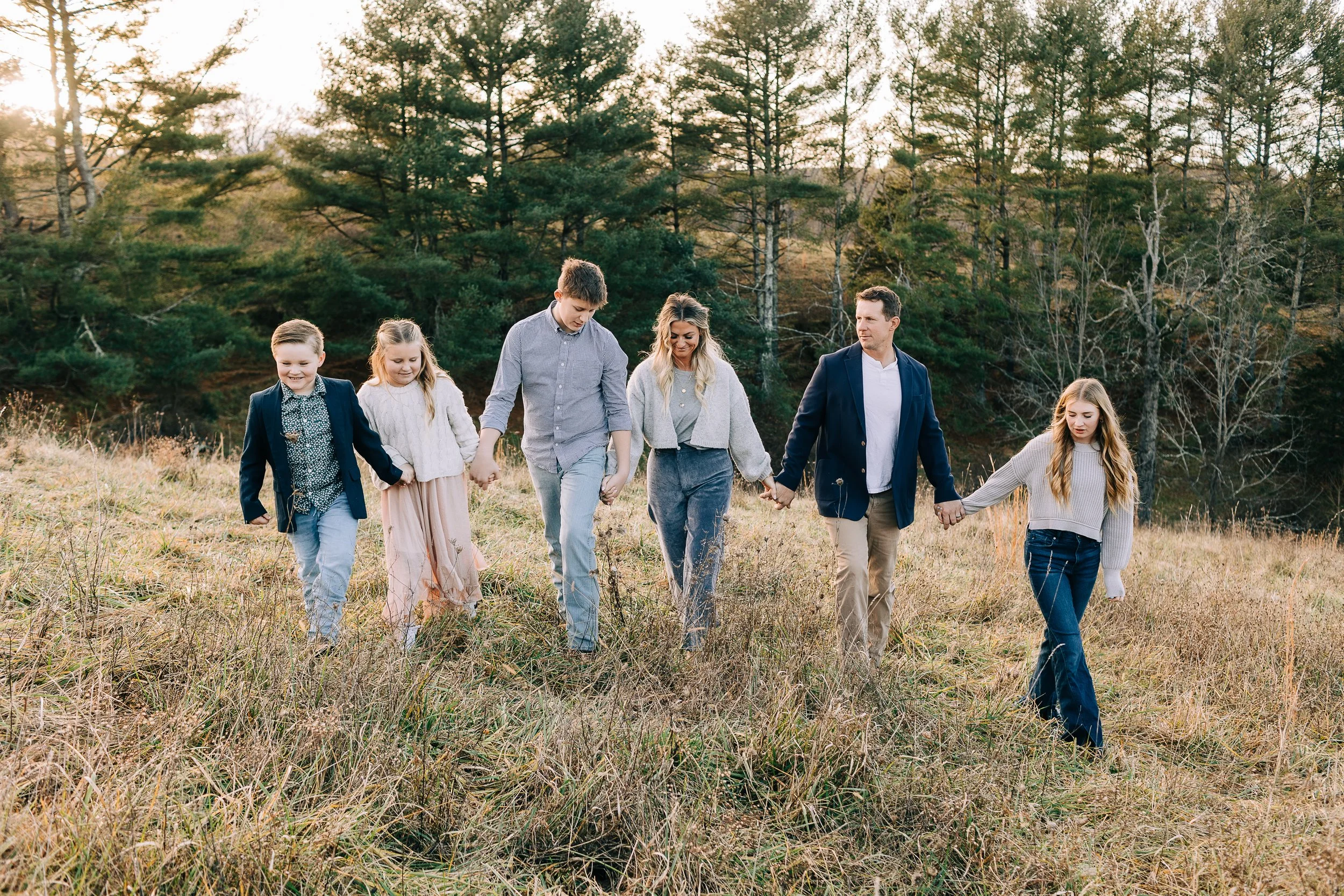 A family of seven holding hands and walking through a grassy field with trees in the background during daytime.