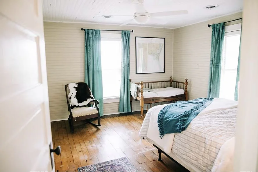 Bedroom with wood flooring, light-colored walls, and two windows with teal curtains. There is a bed with white bedding and a teal throw, a wooden baby crib, a black and white rocking chair, and a framed map on the wall.