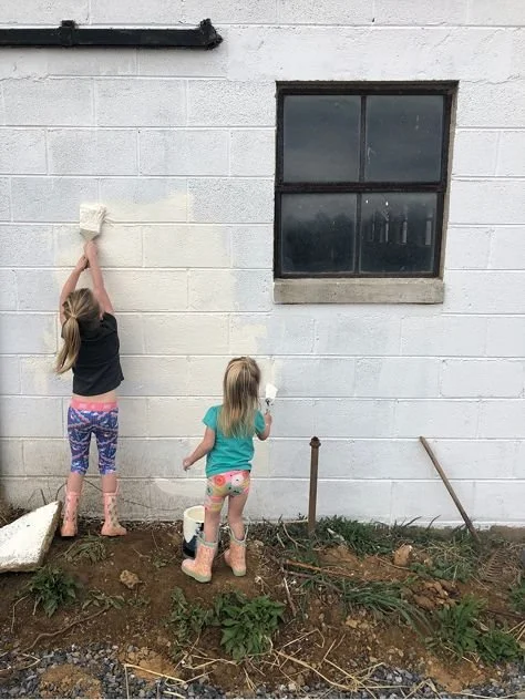 Two young girls paint the exterior wall of a building. One girl uses a roller brush on the wall, while the other holds a small roller and paints near a window.