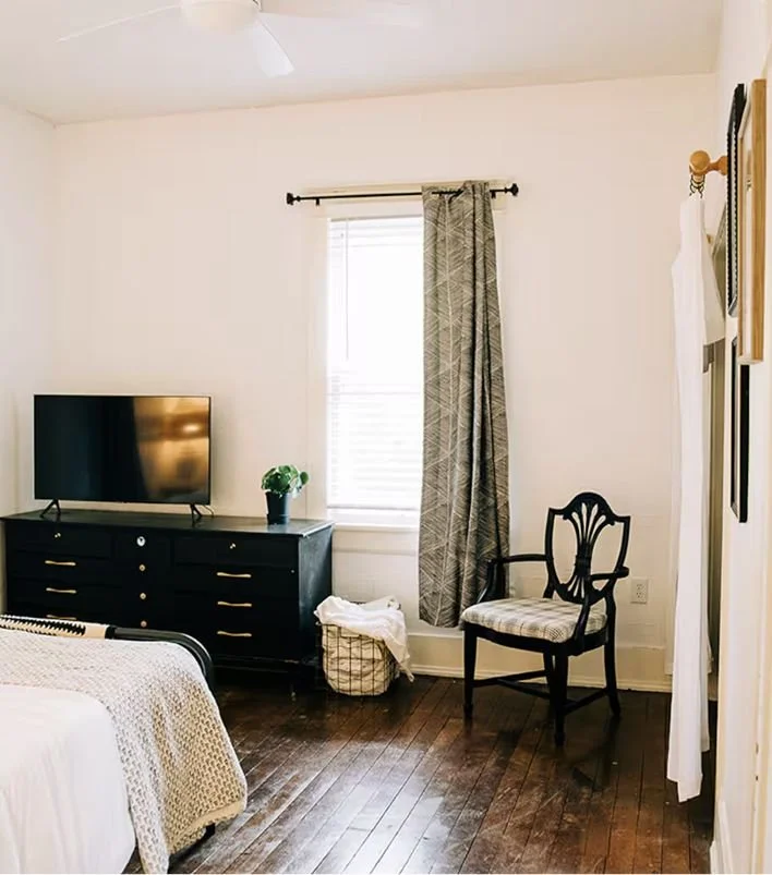 A bedroom with a flat-screen TV on a black dresser, a small potted plant, a window with blinds and patterned curtains, an elegant wooden chair with a cushion, and a laundry basket on the hardwood floor.