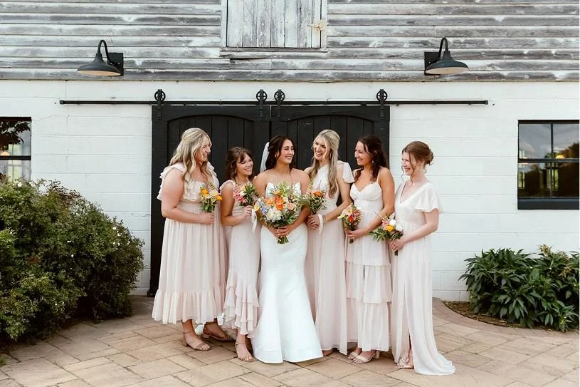 A bride and five bridesmaids standing together outside a rustic barn with sliding black doors, holding bouquets, smiling and looking at each other.