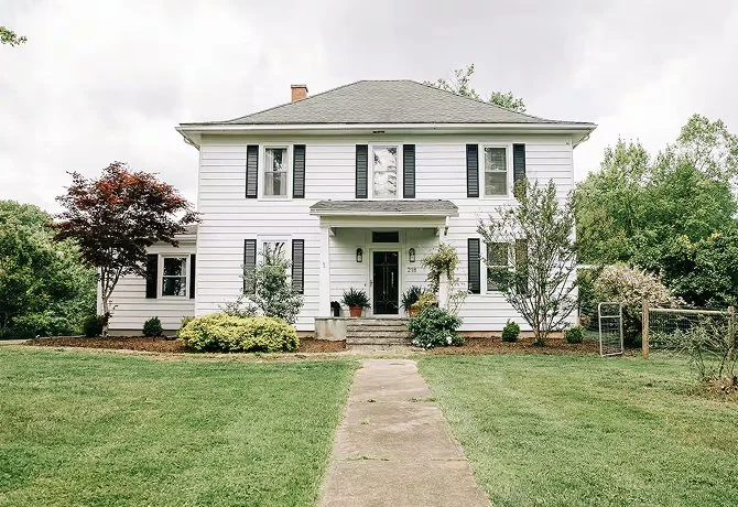 A two-story white house with black shutters, a small front porch, and a gray roof, surrounded by a well-maintained lawn and trees.