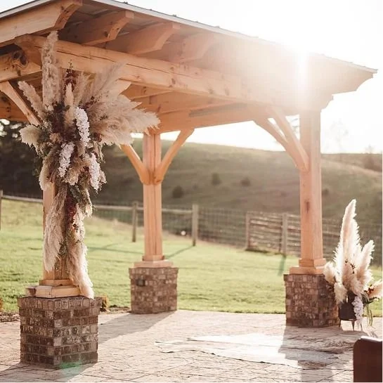 Wooden outdoor pavilion with brick supports decorated with large white pampas grass arrangements, set in a grassy field with a fence and rolling hills in the background, illuminated by sunlight.