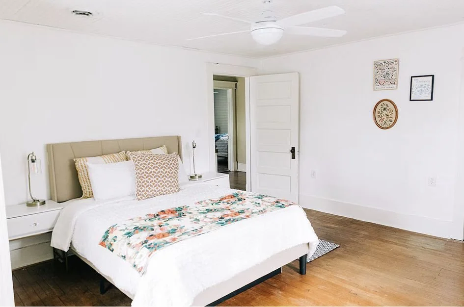 Freshly made bedroom with a large bed, beige headboard, white bedding, floral throw blanket, two white nightstands, slim lamps, white wall with four framed wall art pieces, hardwood floor, and an open door leading to another room.