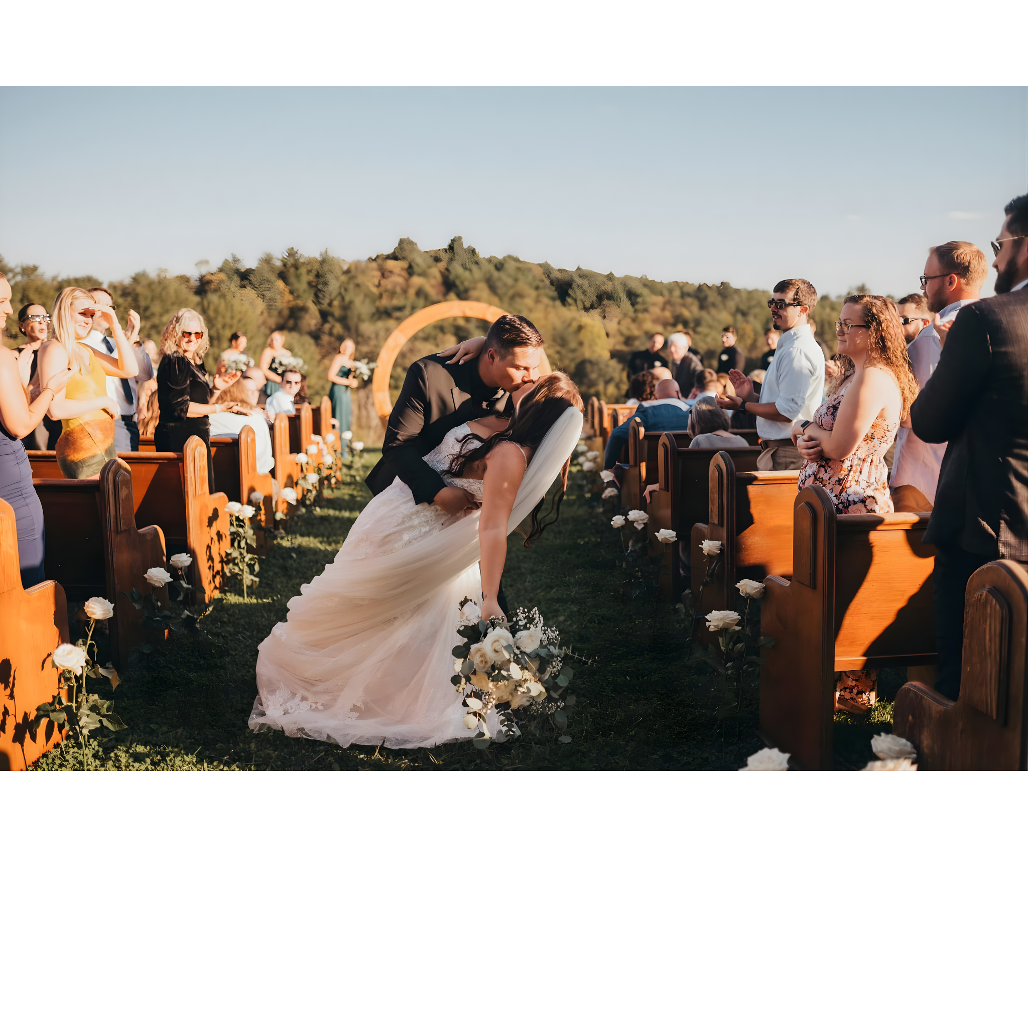 Bride and groom kissing during outdoor wedding ceremony, guests watching on, sunny day with blue sky and trees in background.