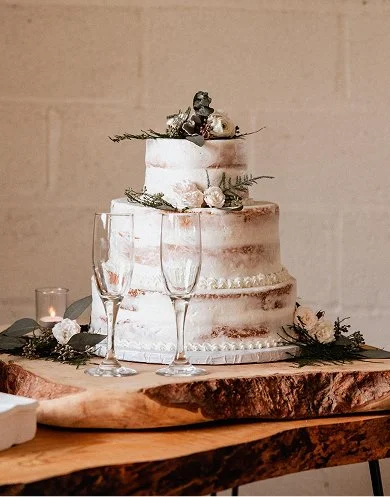 A three-tier wedding cake decorated with greenery, white flowers, and a small animal figurine on top, placed on a wooden table with two empty champagne glasses and lit candles in the background.