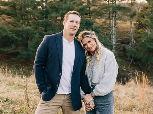 A man and a woman standing outdoors in a natural setting with trees and grass, holding hands, smiling, and looking at the camera.