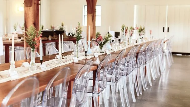 Long dining table set with white candles, flowers, and white tableware in a bright, rustic room with wooden beams.