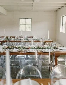 A dining setup with multiple tables adorned with white flowers and glassware in a bright, minimalist room with white walls and large windows.