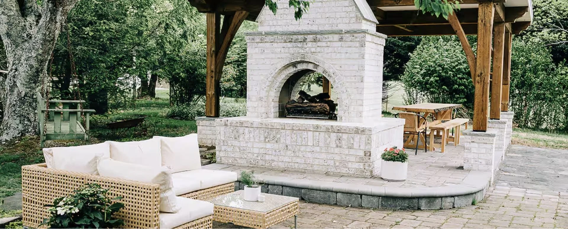Outdoor patio with white brick fireplace, white wicker sofa with cushions, wooden table and chairs, potted plants, surrounded by trees and greenery.