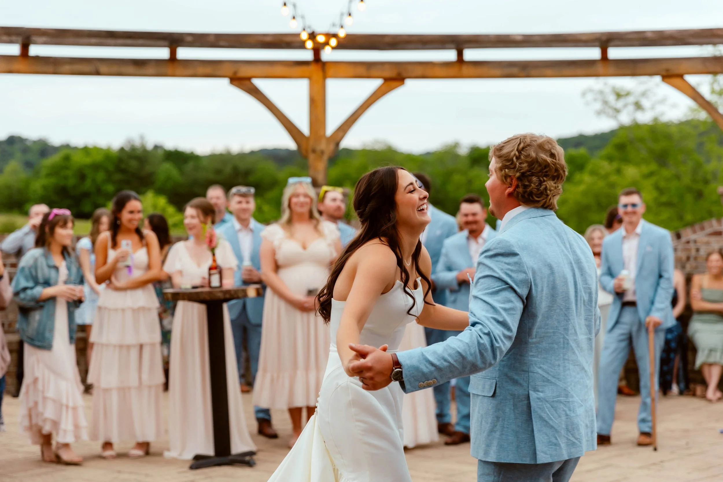 A bride and groom share a dance at their outdoor wedding reception, surrounded by friends and family, with lush green trees and string lights overhead.