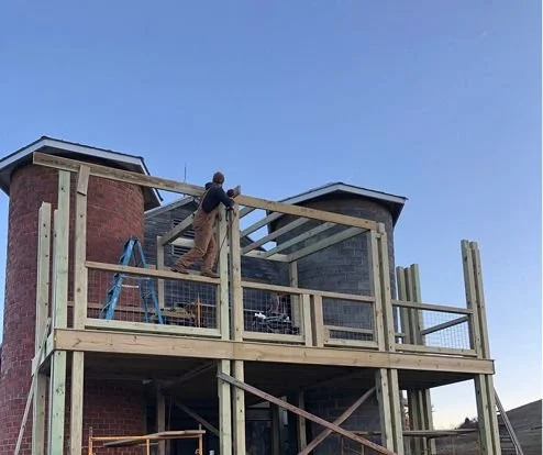 A worker building a wooden deck on the second floor of a brick house, with a ladder and safety railing in place.