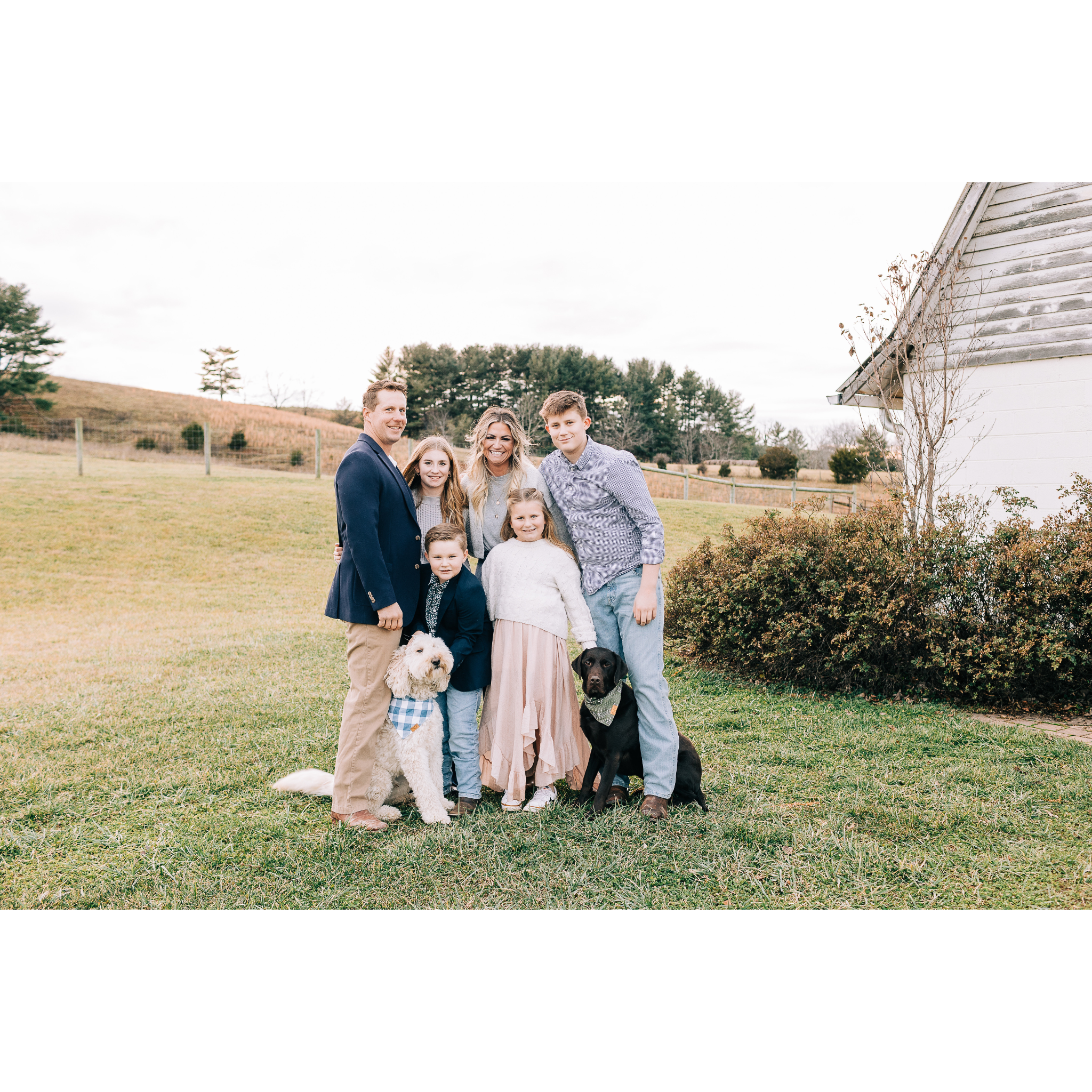 A family of six with two dogs standing on a grassy field outdoors, smiling for a photo, with a rural landscape background.