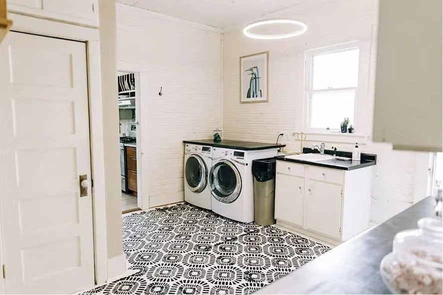 Laundry area with a washing machine, dryer, sink, window, and abstract artwork on the wall in a bright room with patterned black and white floor tiles.