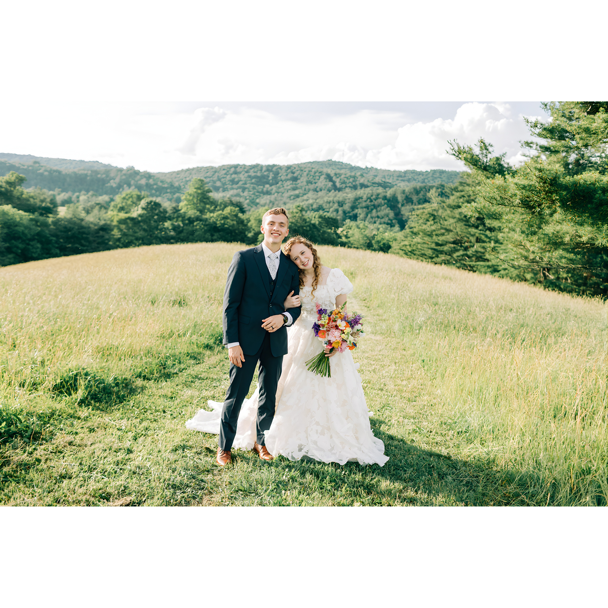 A newlywed couple standing in a grassy field, with the bride leaning her head on the groom's shoulder. The bride is holding a vibrant bouquet of flowers, and the groom is dressed in a dark suit. The background features green trees and rolling hills under a partly cloudy sky.