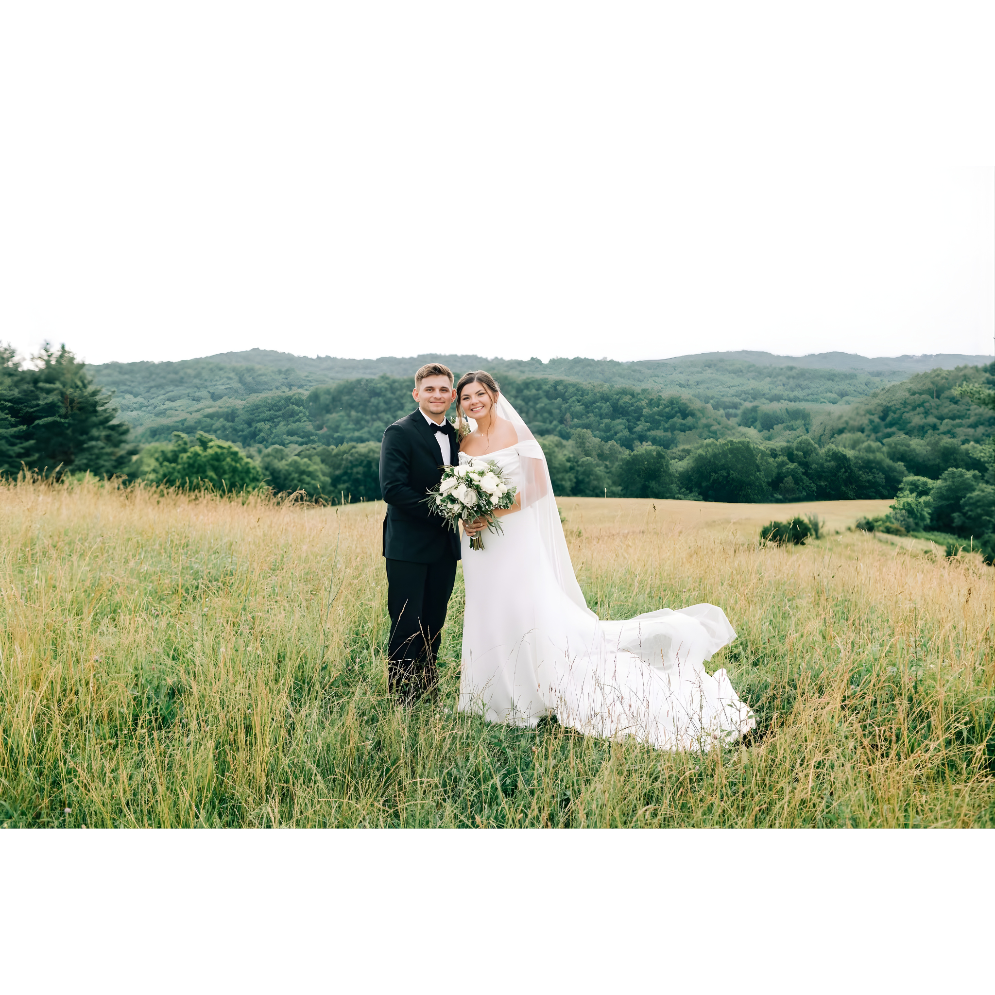 A bride and groom standing together in a grassy field with rolling hills in the background, dressed in wedding attire.