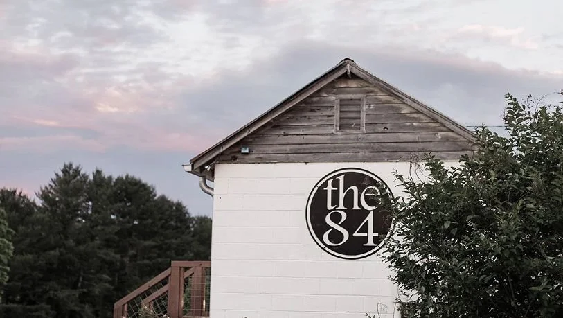 A white building with wooden siding on the upper part and a black circular sign that reads "the 84" on the side. There are trees and a pinkish sky in the background.