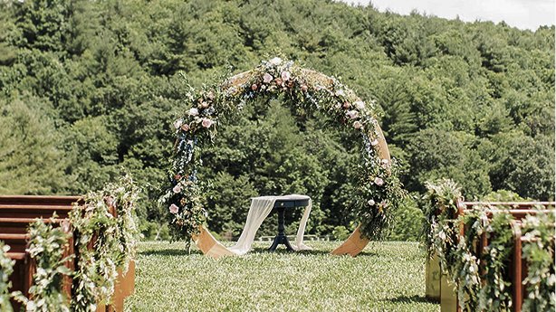 A wedding ceremony setup outdoors with a floral arch and a small table, surrounded by rows of chairs decorated with flowers, on a grassy field with trees in the background.