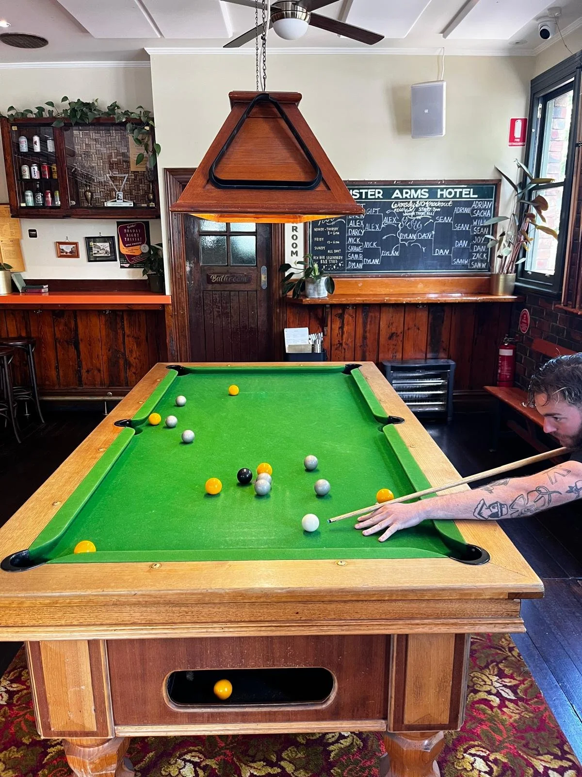 A man plays pool at a wooden table in a pub with a green felt surface, with billiard balls scattered on the table and holding a cue stick, in a room with wooden paneling and a chalkboard sign in the background.
