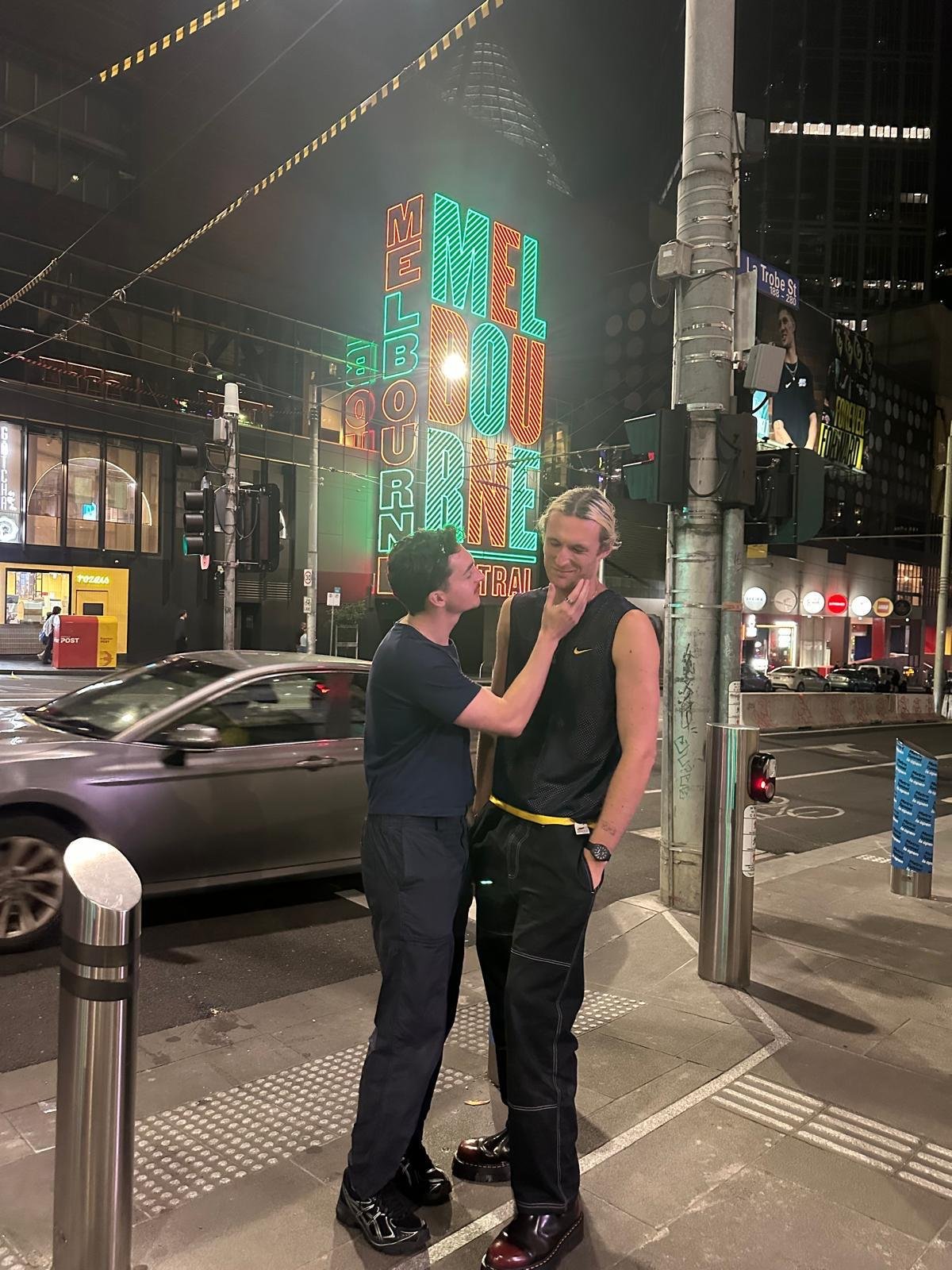 Two young men standing on a city sidewalk at night, one touching the other's chin, both smiling. Behind them, a brightly lit neon sign reads 'Melbourne' and 'CBD' with colorful lights, and a moving car passes by.