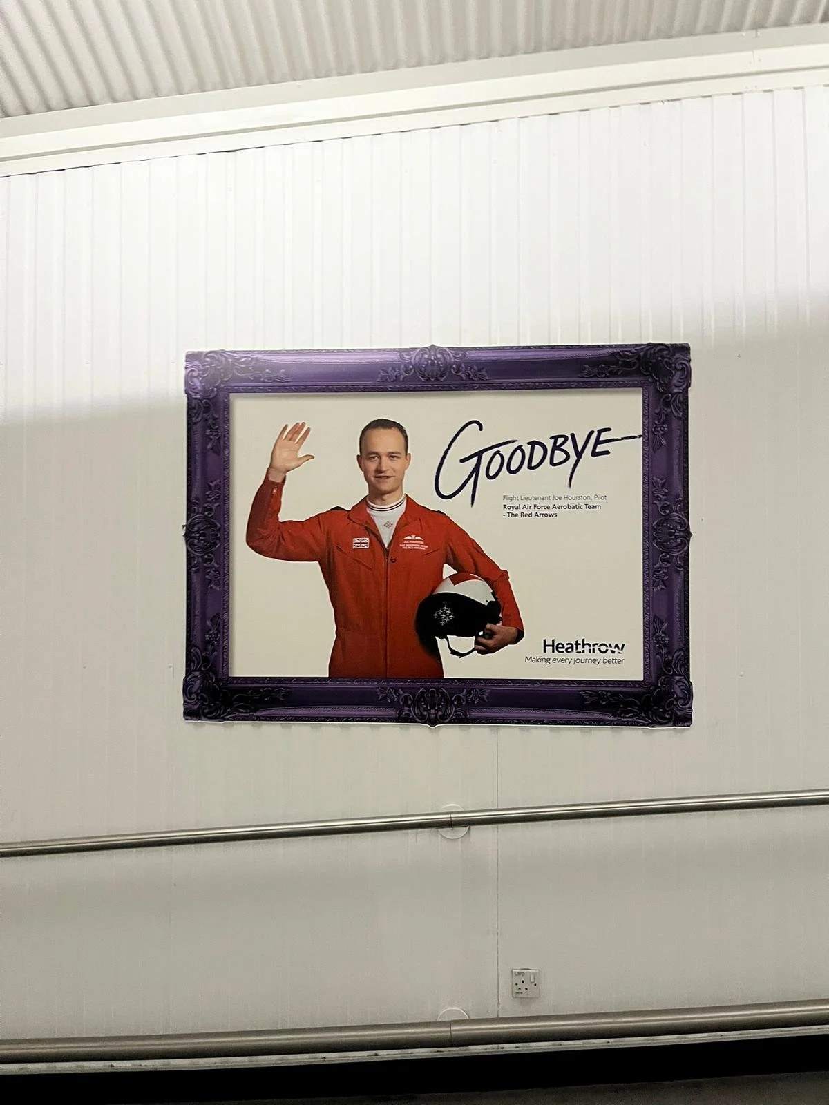 A framed poster shows a man in a red pilot jacket holding a helmet, waving, with the word "Goodbye" written across it. The poster includes the Heathrow logo and text about flight Lieutenant Joe Houston, a Royal Air Force pilot with the Red Arrows.