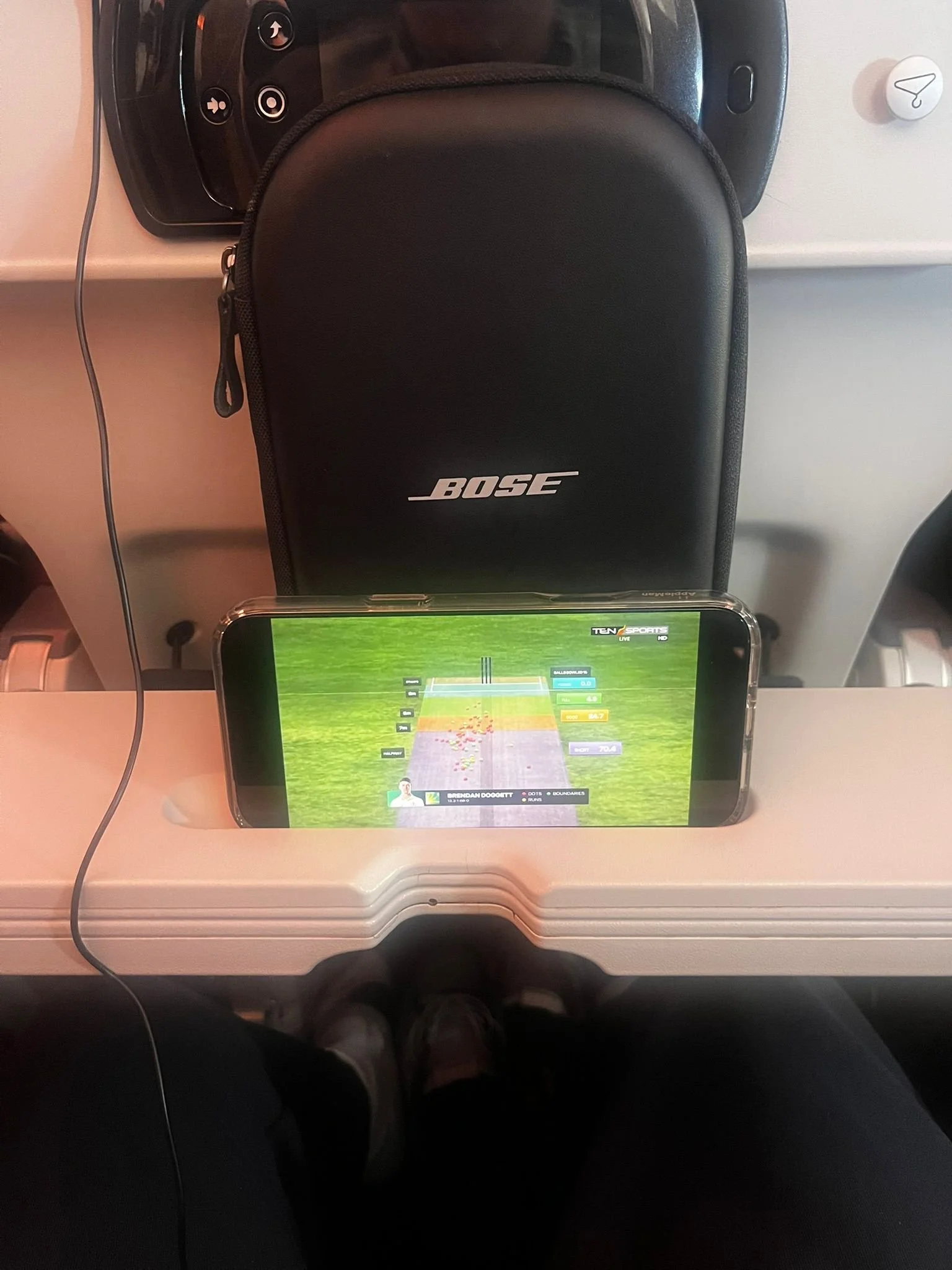 A person's view from a plane seat, showing a smartphone on a foldable tray table displaying a cricket match, with a Bose headphone case and a white surface nearby.
