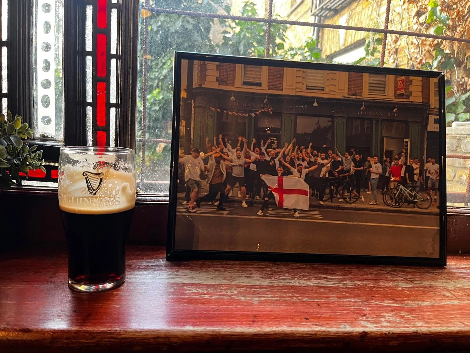 A pint of dark stout beer with foam on top, placed on a wooden surface beside a framed photo of a group of people celebrating on a city street, holding an England flag. The background shows a window with colorful stained glass and greenery outside.