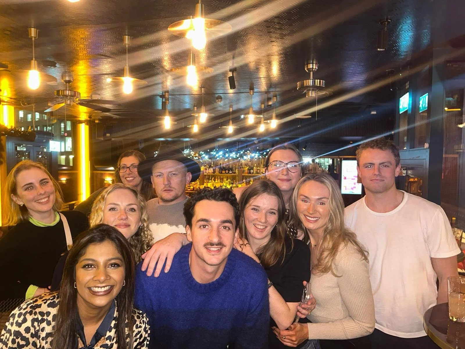 Group of friends gathered in a bar or restaurant, smiling and posing for the photo with a bar in the background, dim lighting, and warm accent lights.