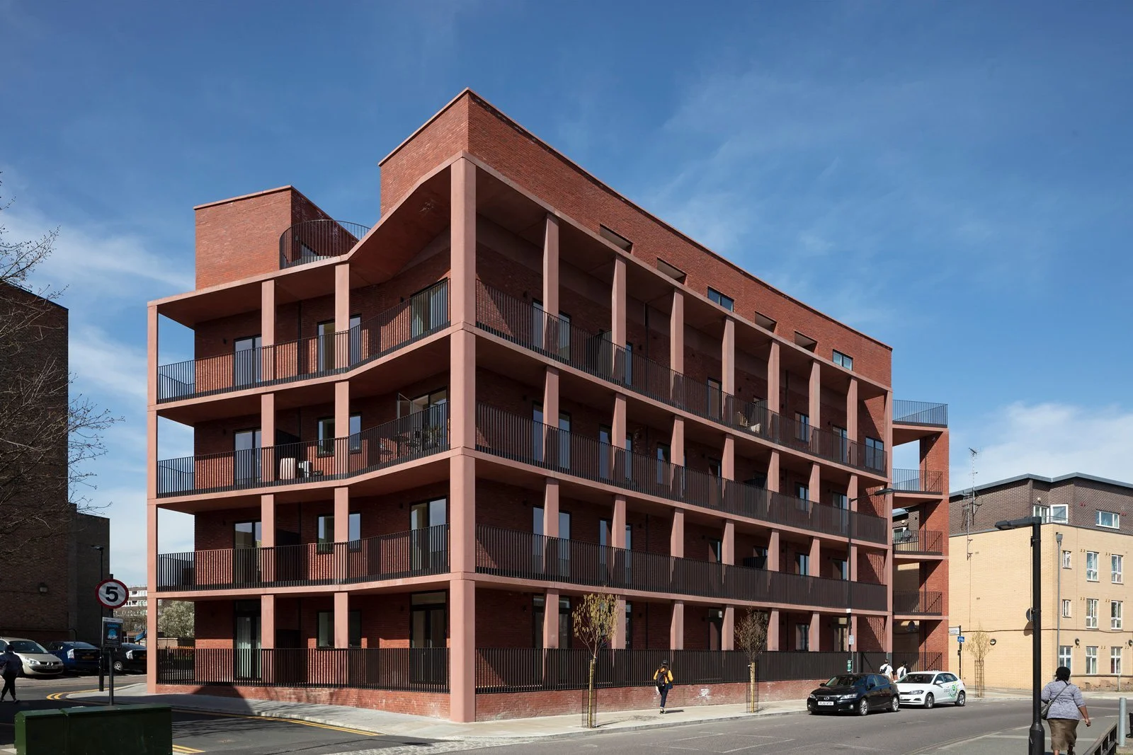 A modern, red-brick multi-story apartment building with multiple balconies and railings, situated on a city street with cars and pedestrians.