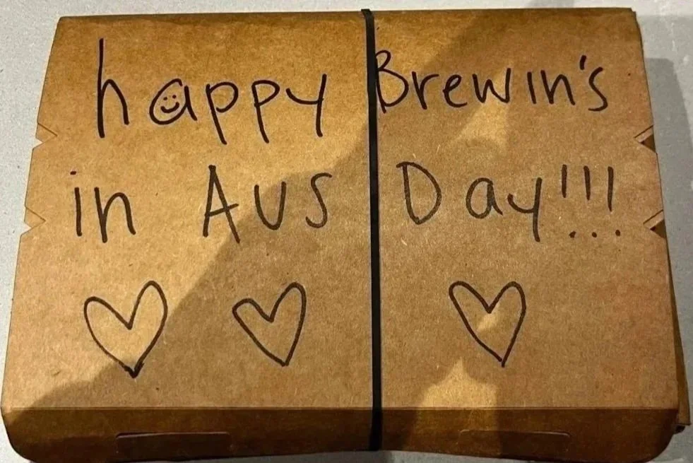 Brown paper takeout box with handwritten message celebrating Brewin's 'Austin's Day' featuring hearts and a smiley face in place of the dot over the 'i' in happy.