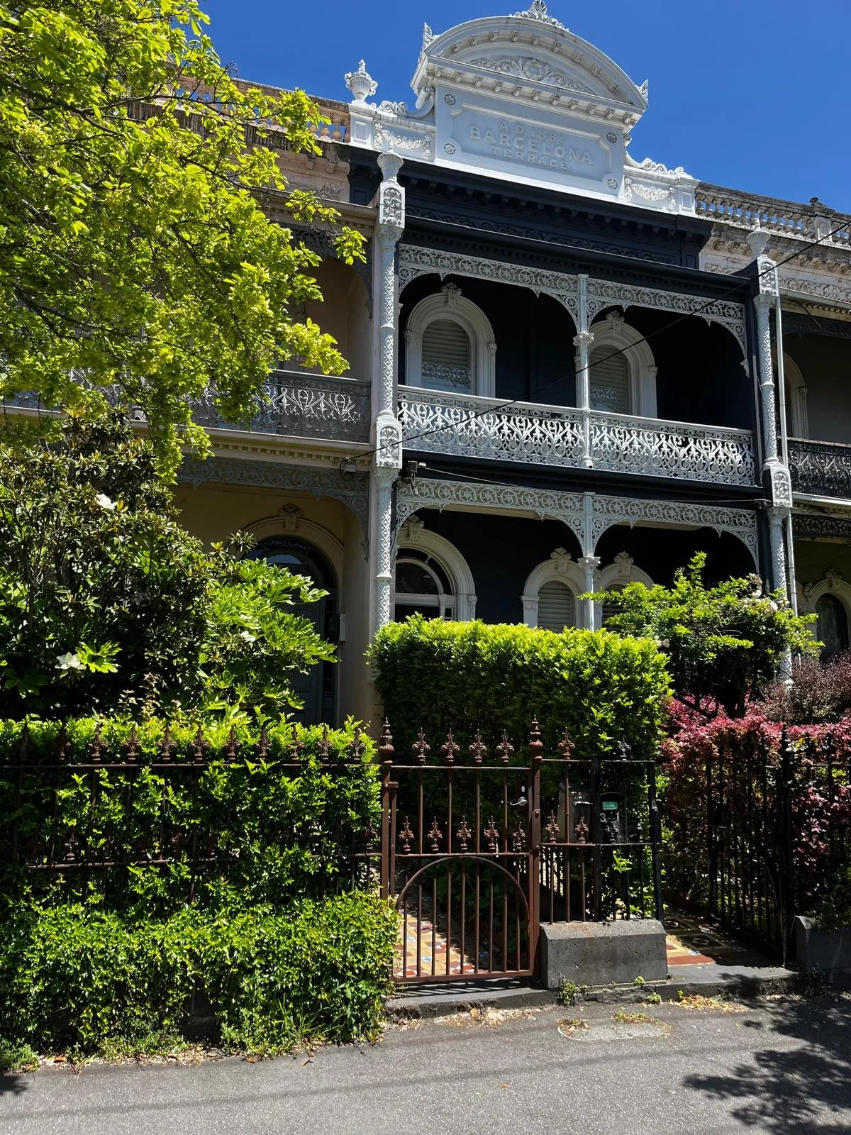 A historic Victorian-style building with ornate black and white ironwork balconies, surrounded by lush green trees and bushes, with a clear blue sky above.
