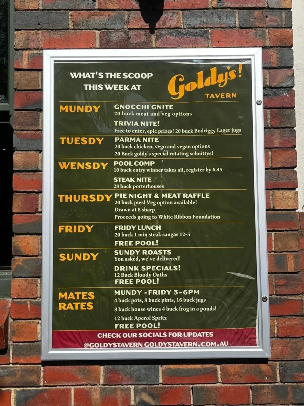 A menu board outside Goldy's Tavern listing weekly events and specials, with a brick wall background.