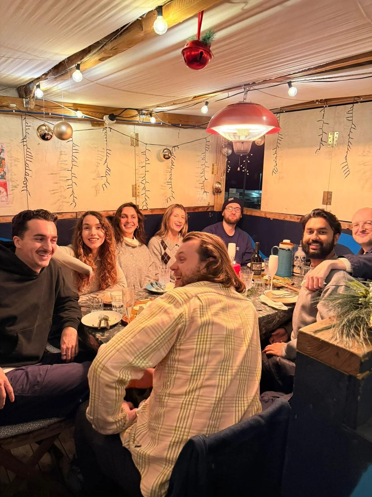 A group of nine people sitting around a dining table, smiling and enjoying a gathering in a decorated, cozy indoor setting.