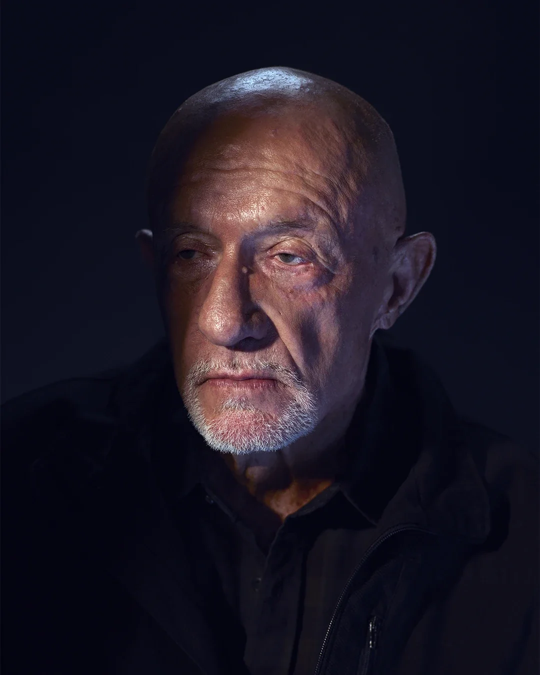 A close-up portrait of an elderly man with a serious expression, bald head, and a white beard, wearing a dark jacket against a dark background.