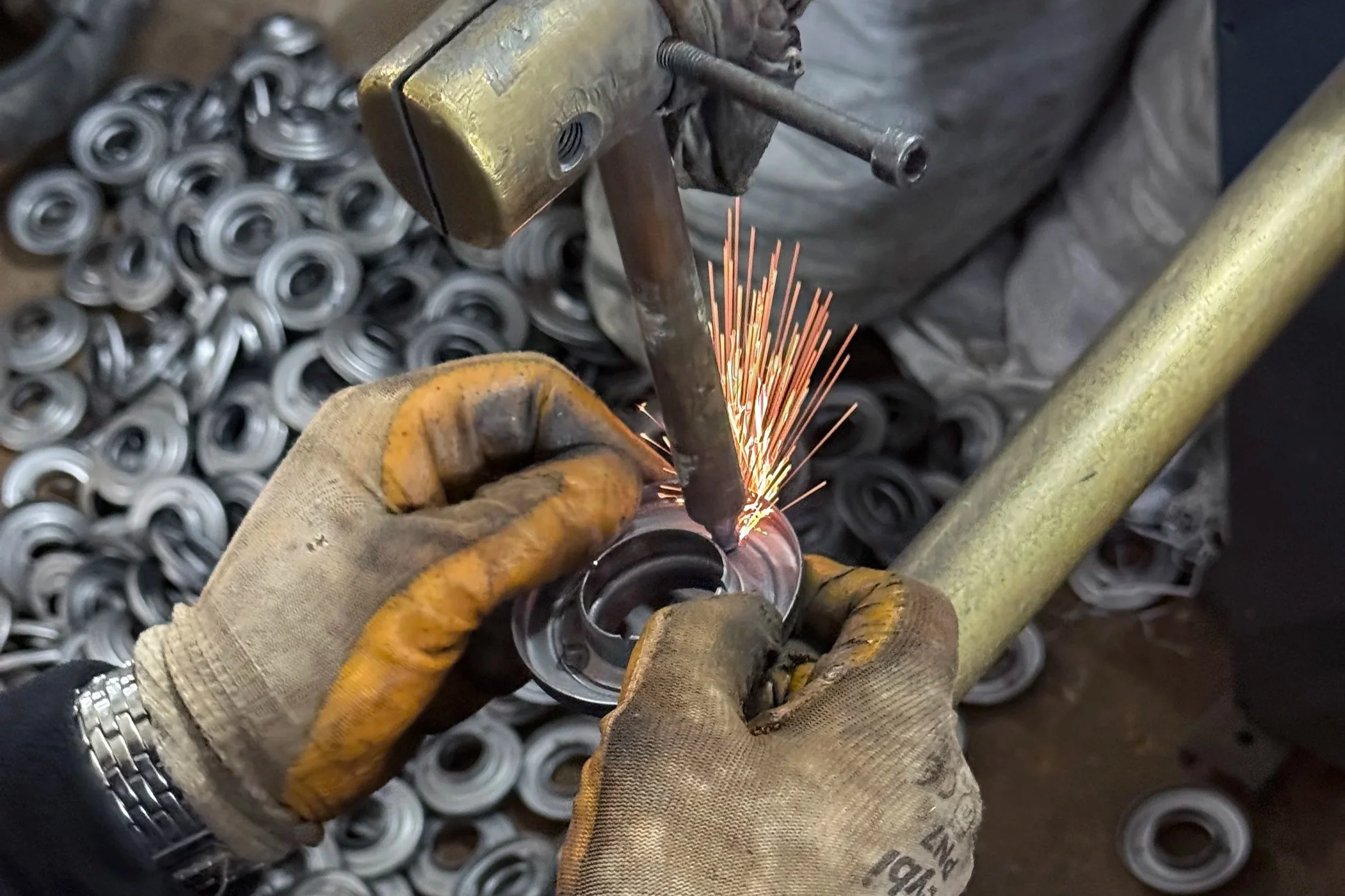 A worker welds a metal part using a welding torch, with sparks flying, in a workshop surrounded by many finished metal rings or components.