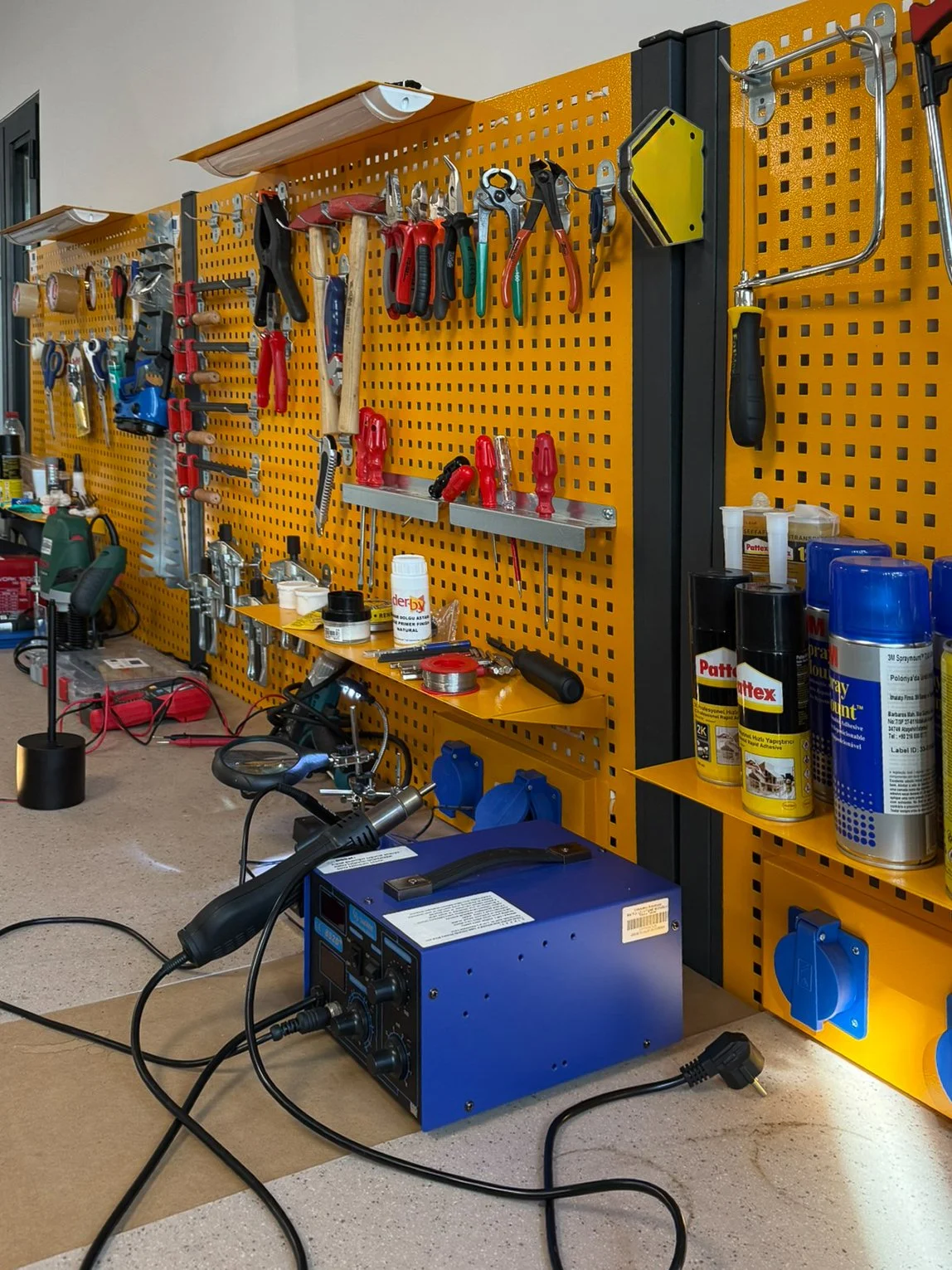 Tools and equipment organized on a yellow pegboard, including hammers, screwdrivers, pliers, a saw, and spray cans, with a blue welding machine on the work surface.