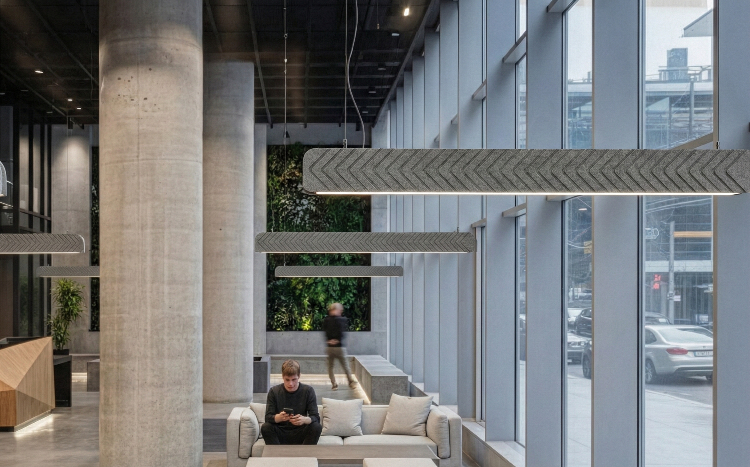 Modern hotel lobby with large windows, concrete pillars, and hanging pendant lights. A man is sitting on a sofa looking at his phone, and another person is walking in the background.