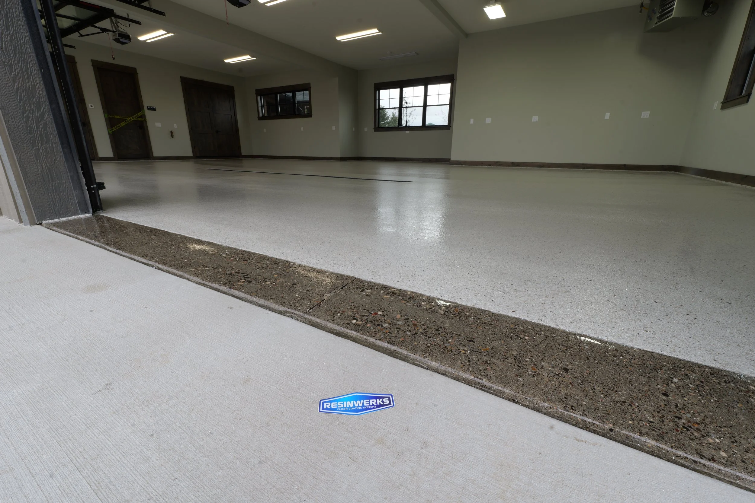 Empty garage with freshly coated epoxy floor, visible resinwerks sticker near the entrance, and windows on the far wall.