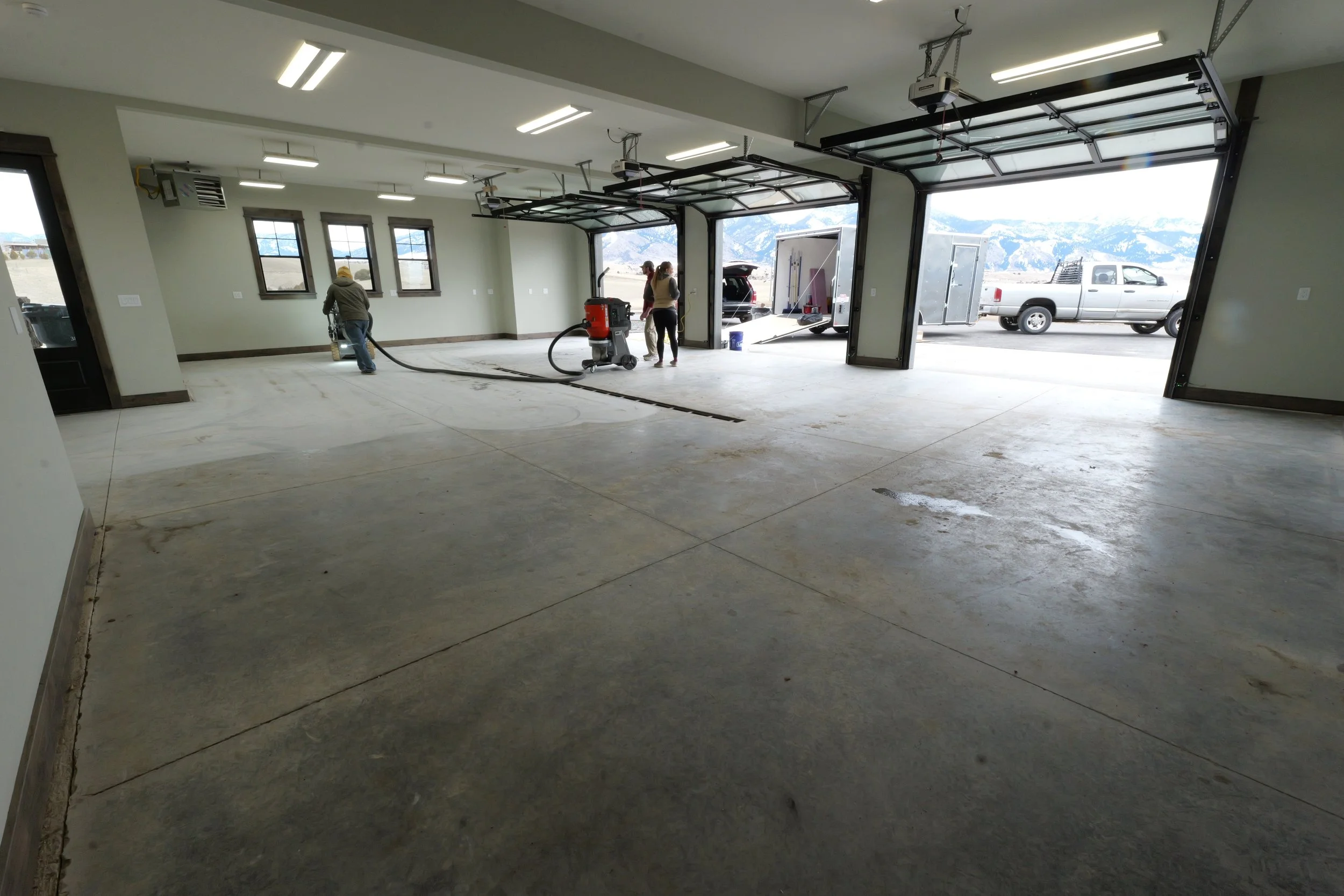 Inside a large garage with open doors, three workers are cleaning the concrete floor with a vacuum or squeegee, and there is a pickup truck parked outside with a trailer. Mountains are visible in the background.